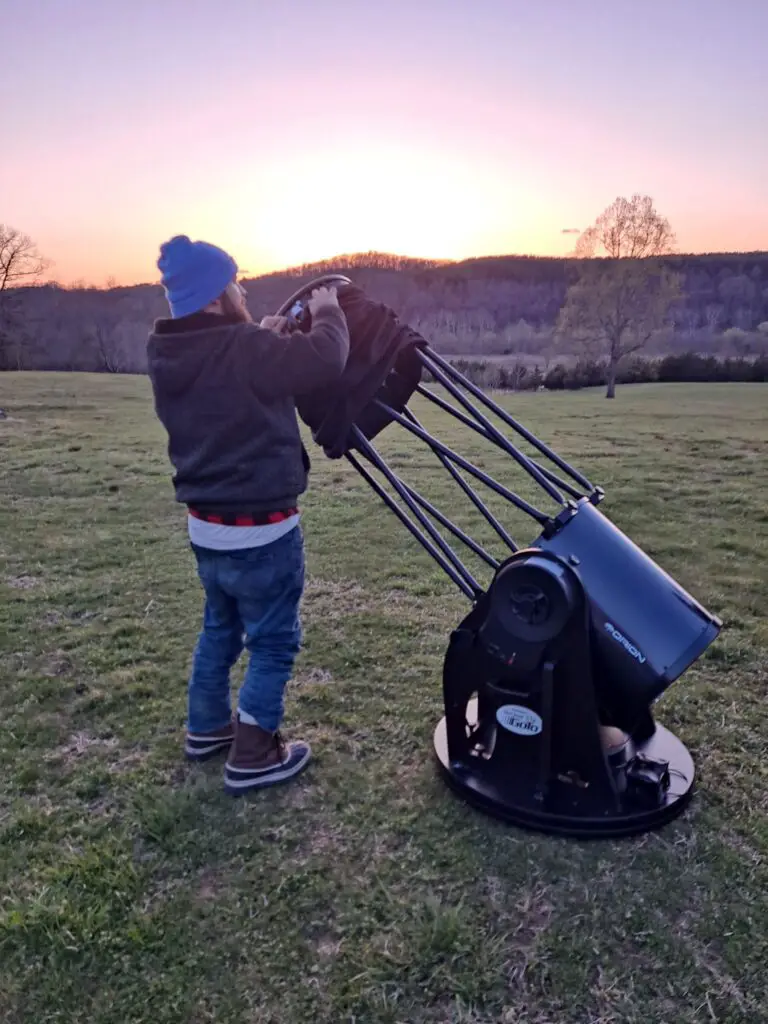 A man standing assembling a large telescope outside in front of a sunset